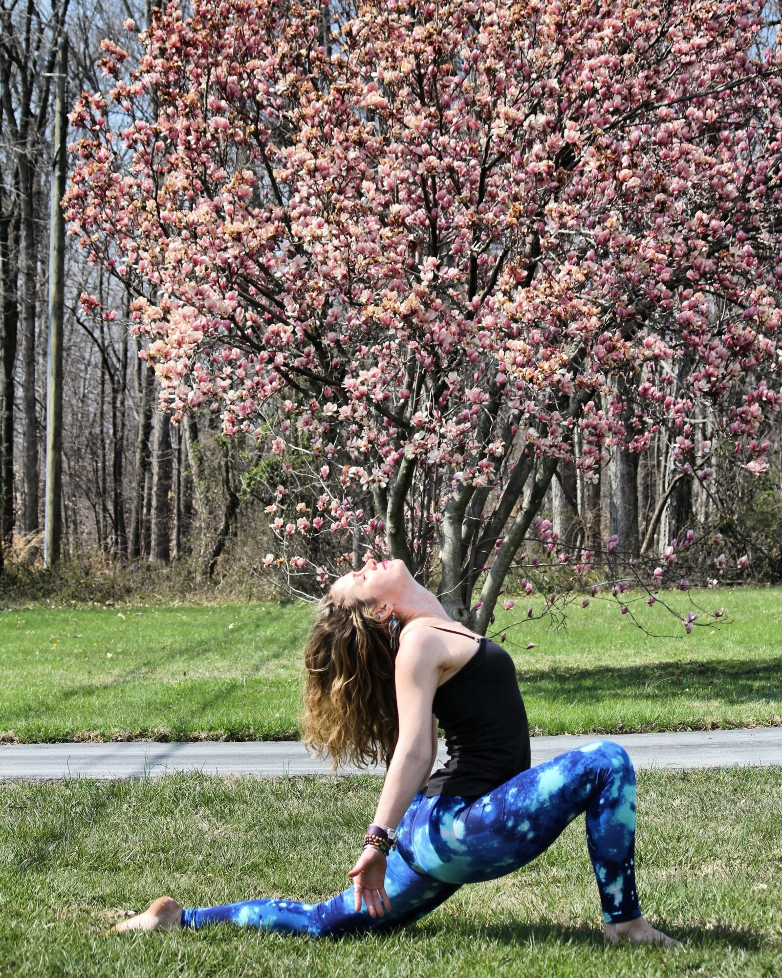 Person practicing yoga outdoors on grass with pink flowering tree and forest in background.