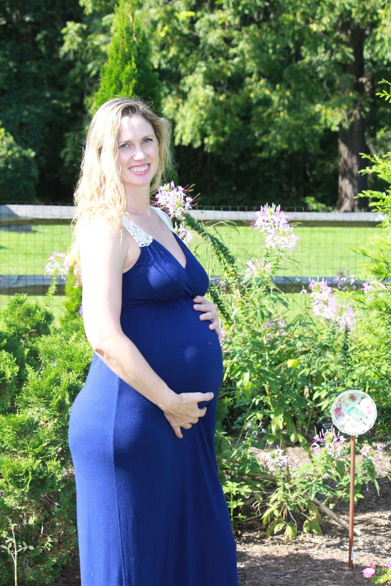 Woman in a blue dress standing outdoors with greenery and flowers, holding her hands in front of her, smiling.