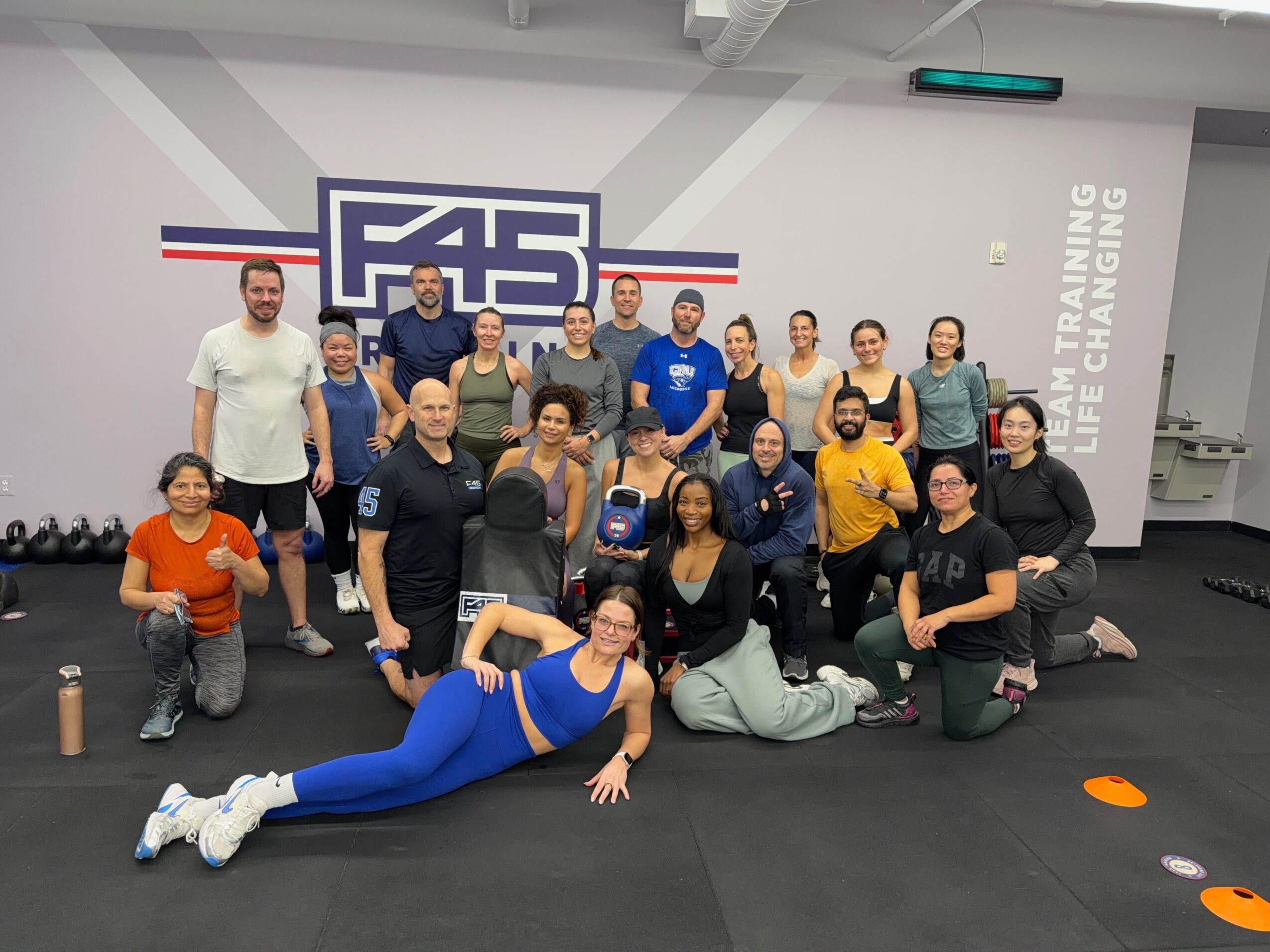 Group of 20 people in a fitness studio, some standing and some sitting or lying on the floor, smiling at the camera.