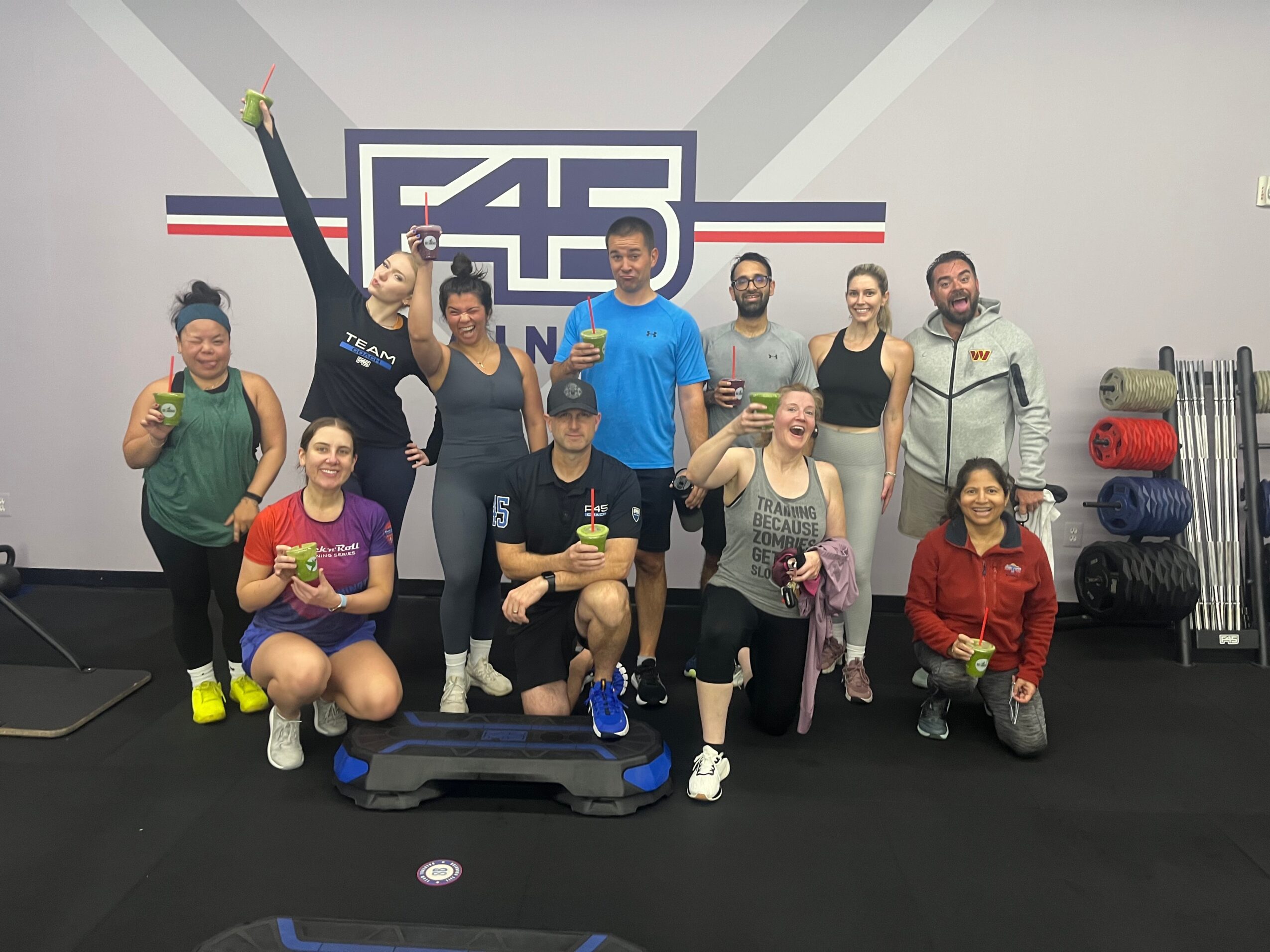 Group of ten people in a gym, some holding medals, standing and kneeling, with fitness equipment and a logo in the background.