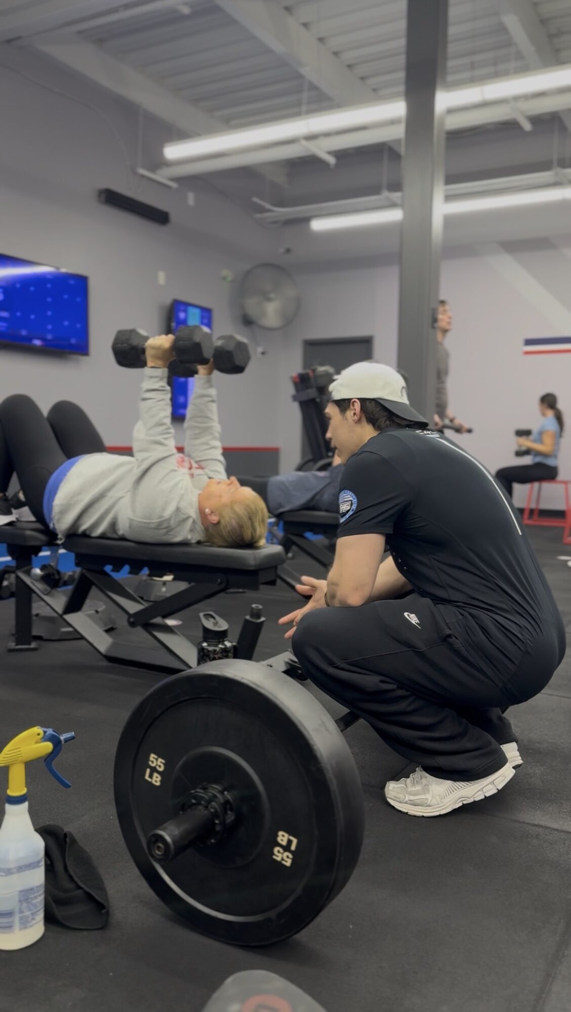 Person lying on a bench press with weights, trainer kneeling beside in a gym setting.