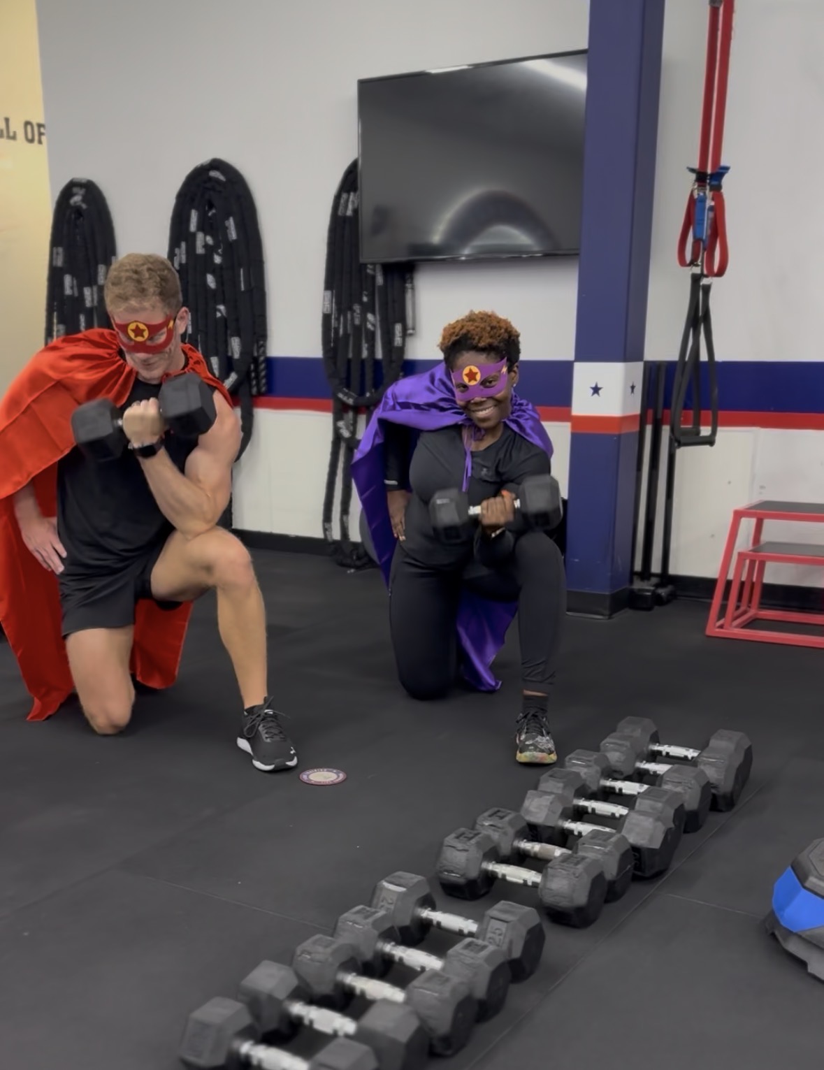 Two children dressed as superheroes with capes and masks in a gym, kneeling near a row of dumbbells, smiling.