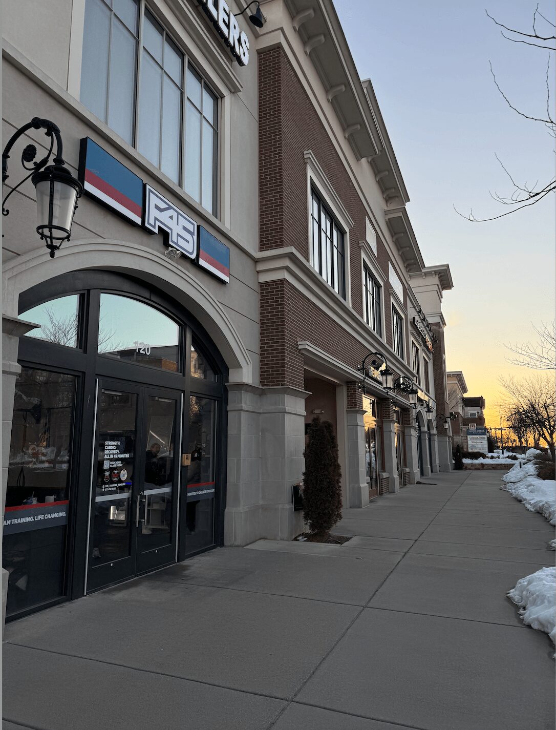 Street view of a building with a sign, entrance, and sidewalk, with snow on the ground and trees in the background.