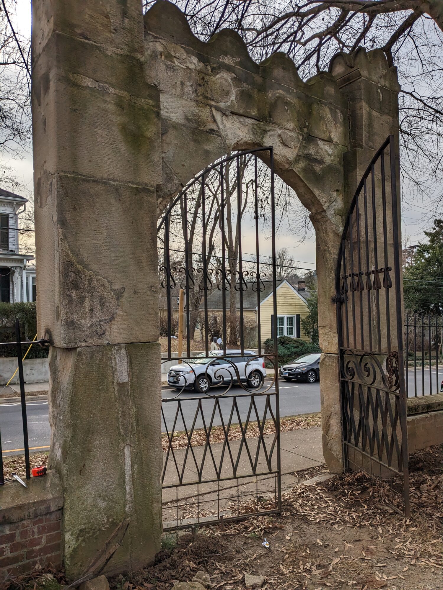 Stone archway with iron gate, street with parked cars and houses in background, leafless trees, cloudy sky.