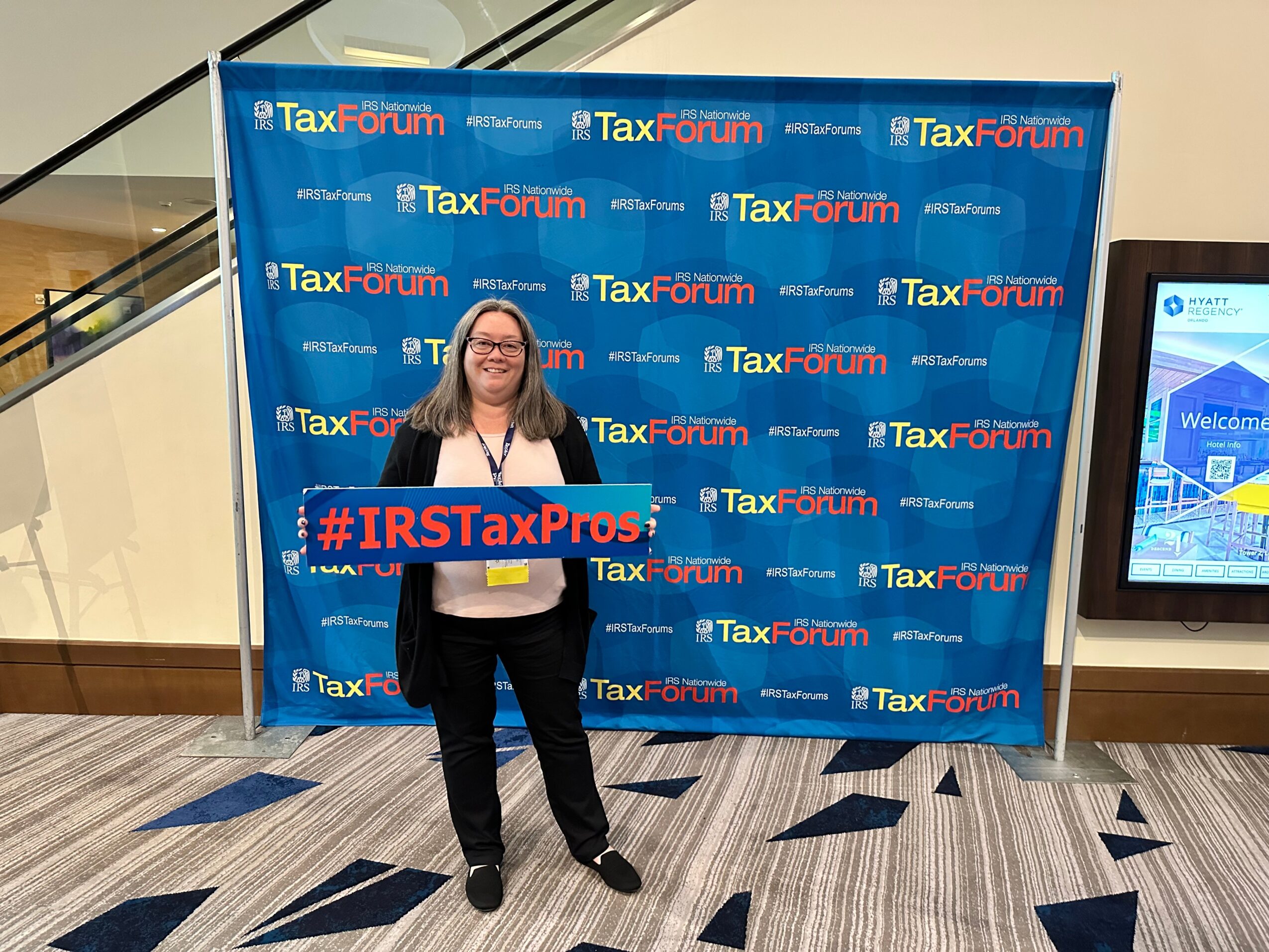 Woman standing in front of a blue Tax Forum backdrop holding a sign, smiling, in a conference setting.