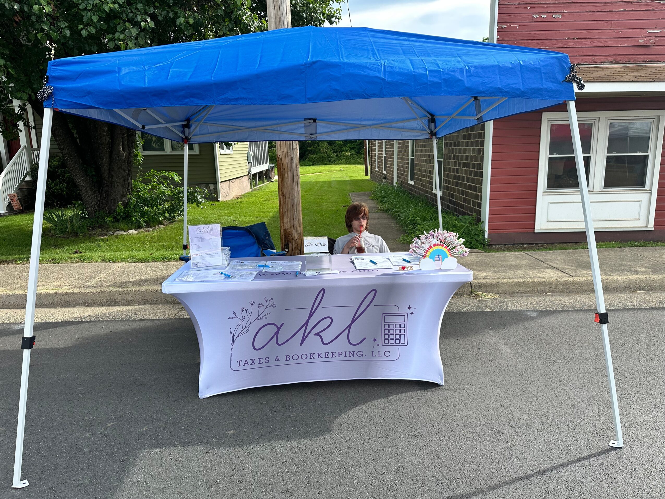 A table with a white tablecloth displaying the logo and name of a cake and bookkeeping business, under a blue canopy on a street.