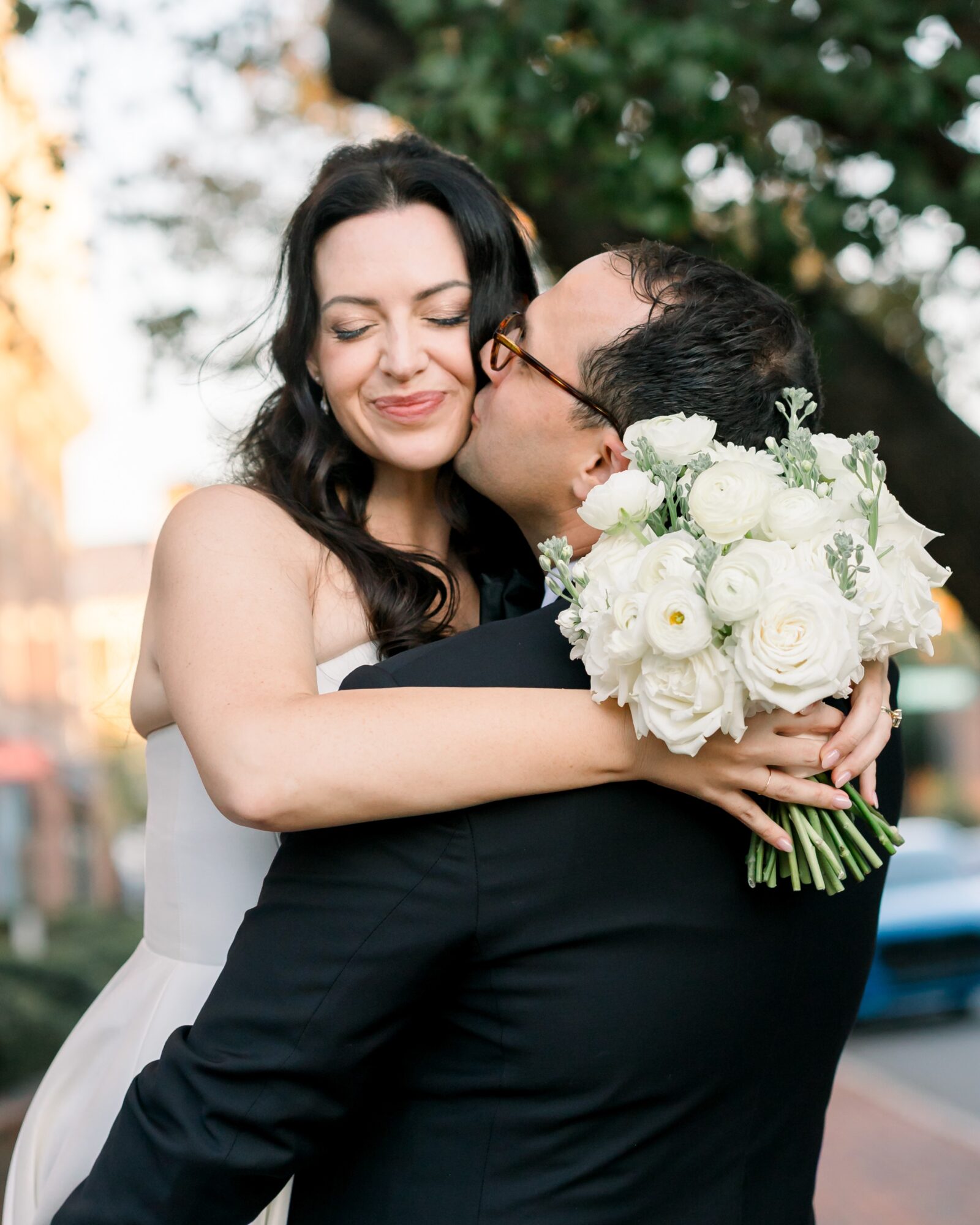A woman with dark hair embraces a man holding a bouquet of white flowers outdoors, smiling with eyes closed.