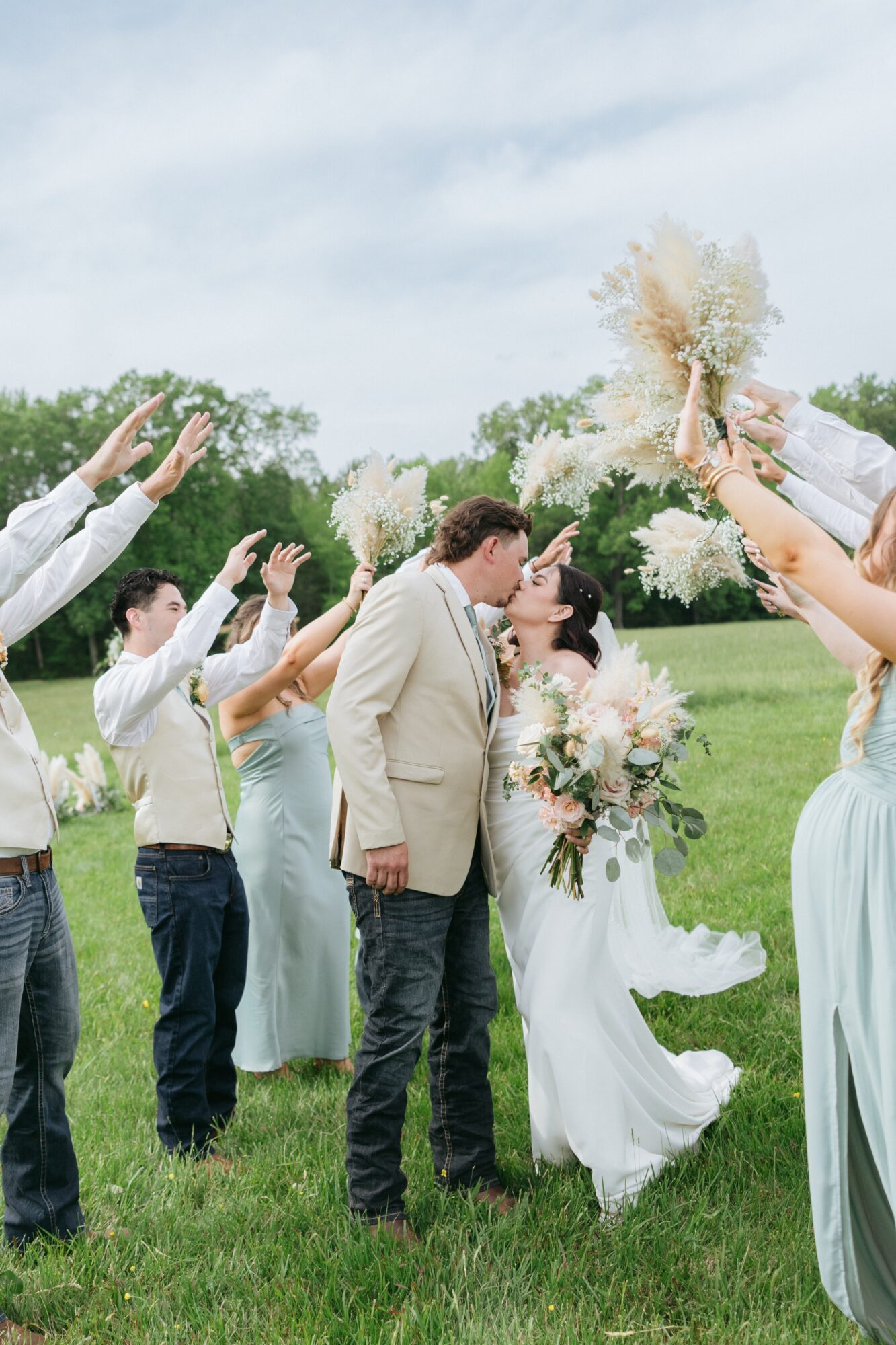 A wedding couple kissing outdoors with friends raising bouquets in celebration, green grass and trees in background, cloudy sky.