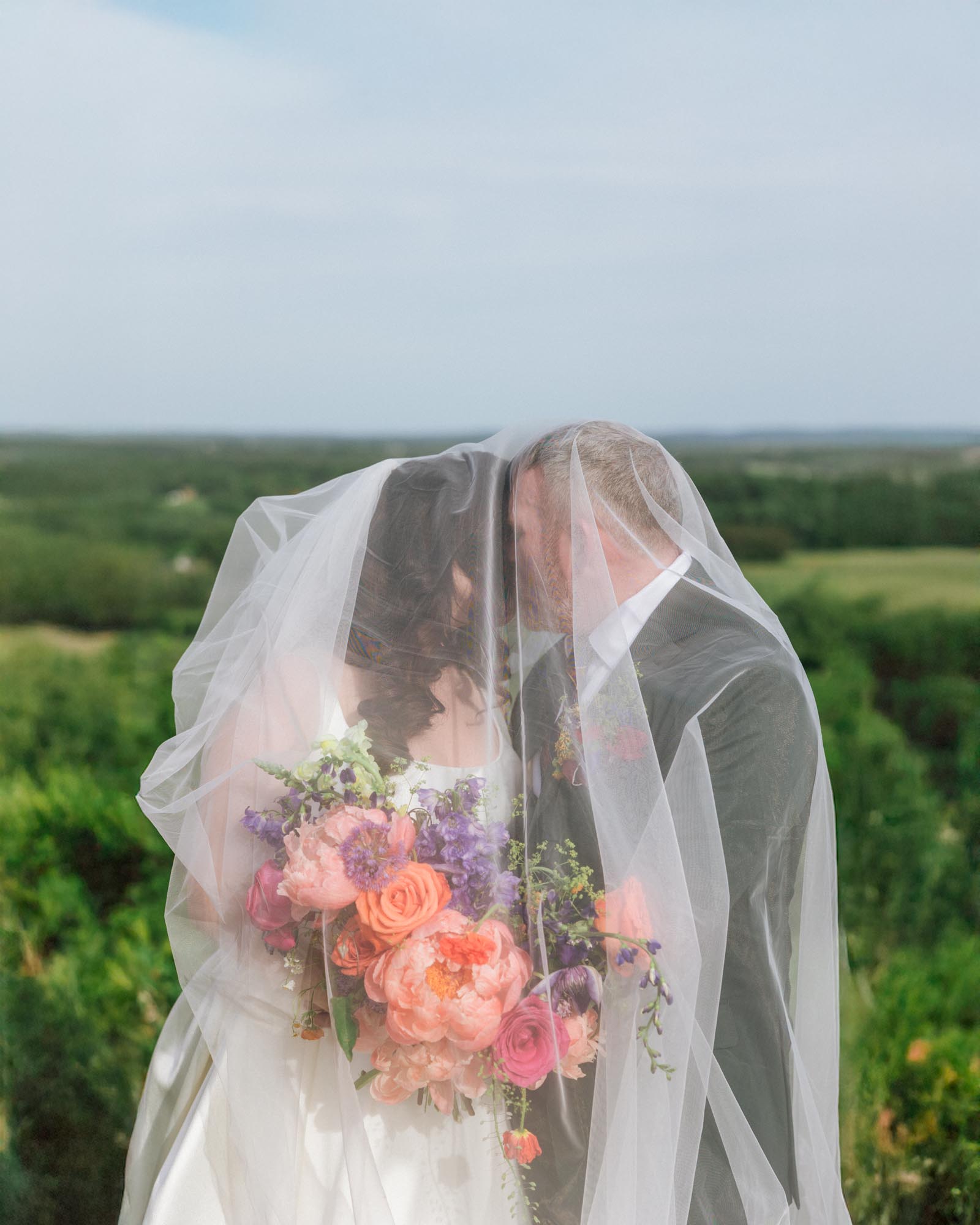 Bride and groom under veil holding a colorful bouquet outdoors, with green landscape background.