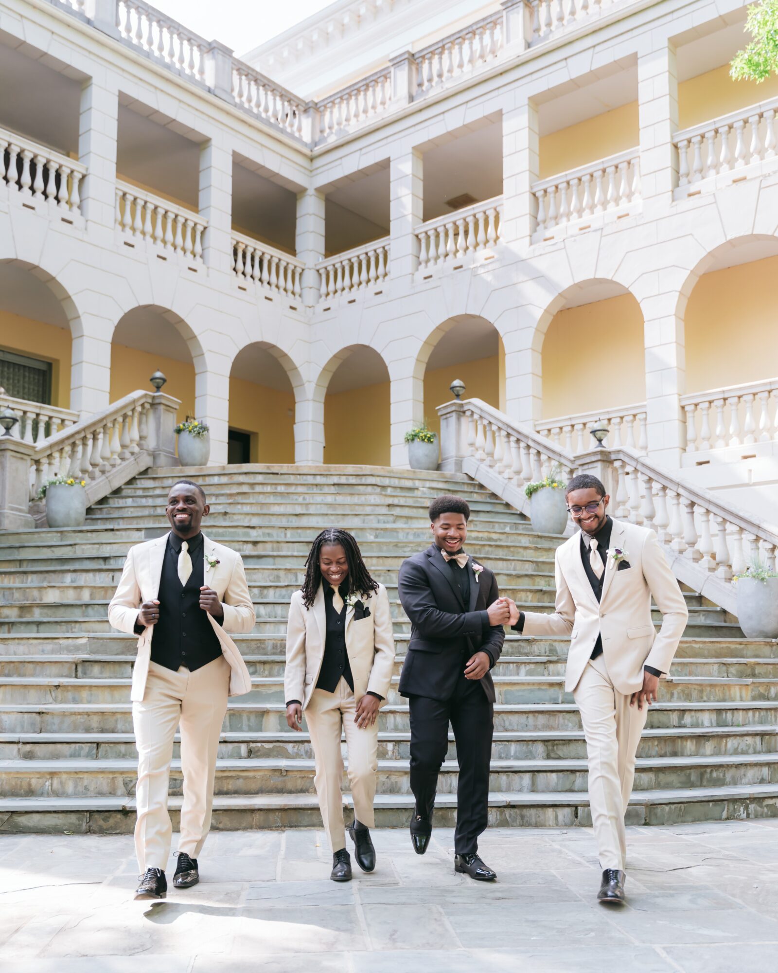 Four people in formal attire walking down stairs outside a building with arches and balconies, smiling and holding hands.