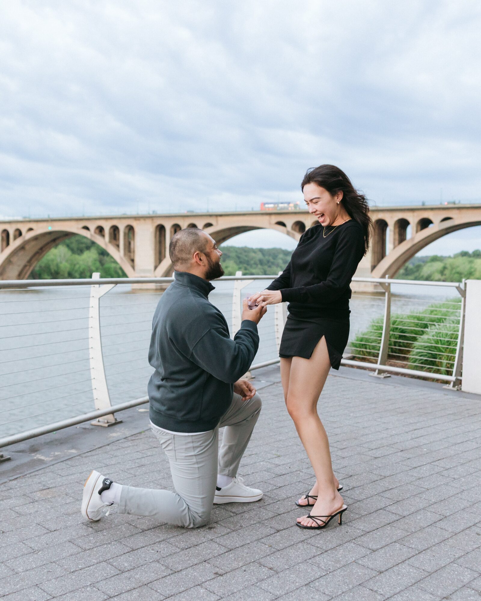 A man kneeling on one knee proposing to a woman on a bridge with a river and arches in the background.