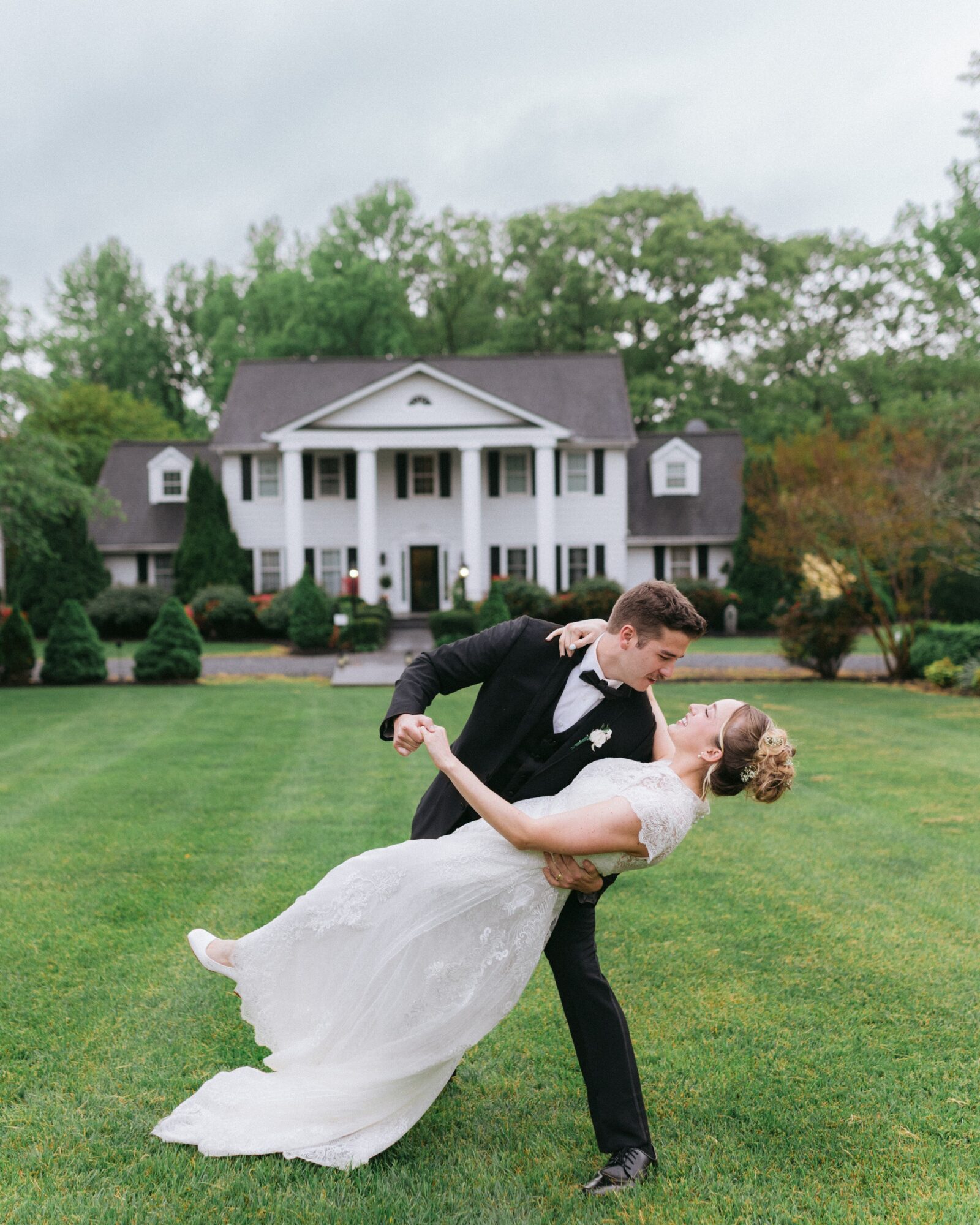 A couple in wedding attire dancing on a lawn in front of a large house with trees in the background.