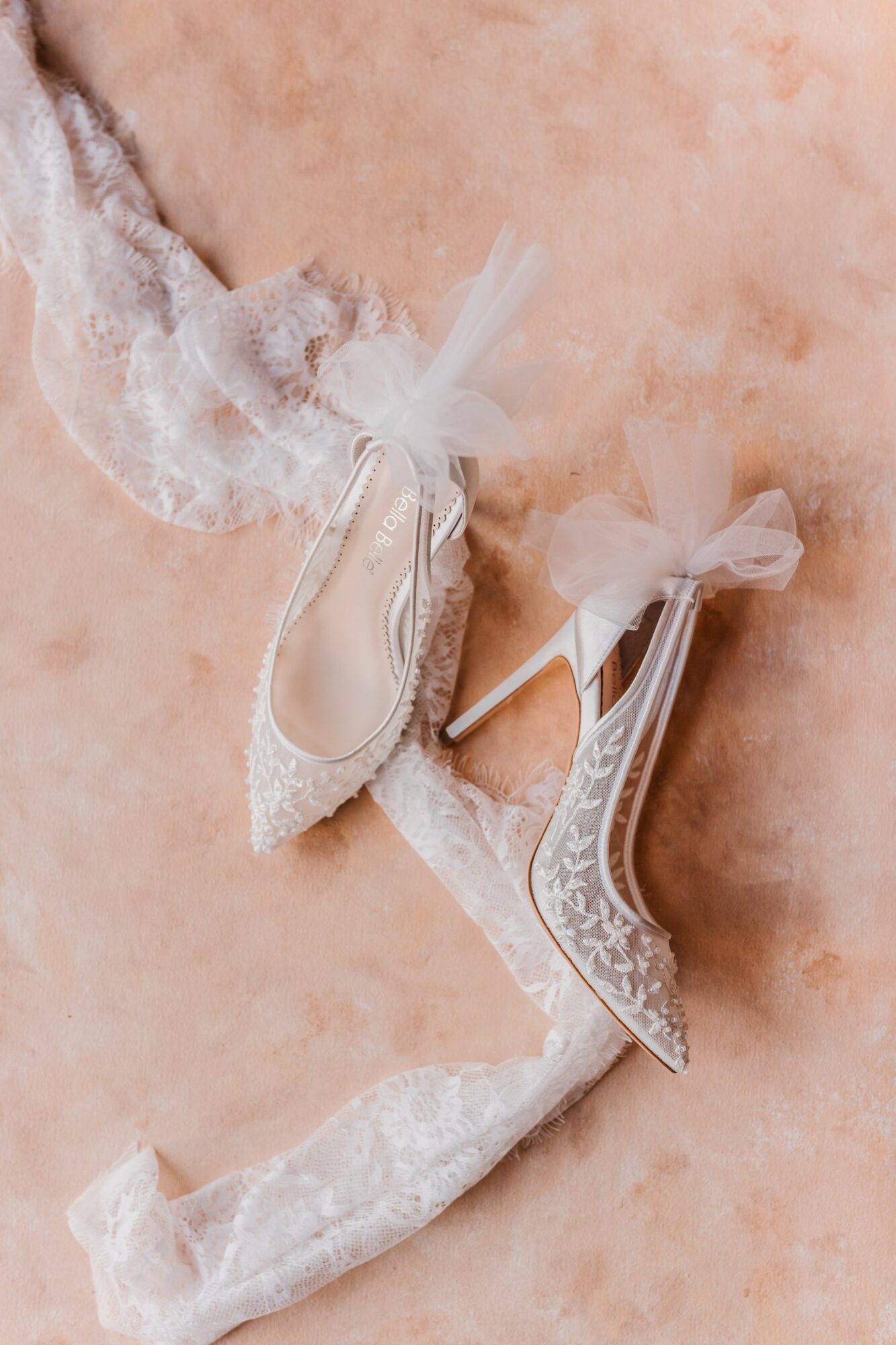 Pair of white lace high-heeled shoes with tulle bows, placed on a textured surface.