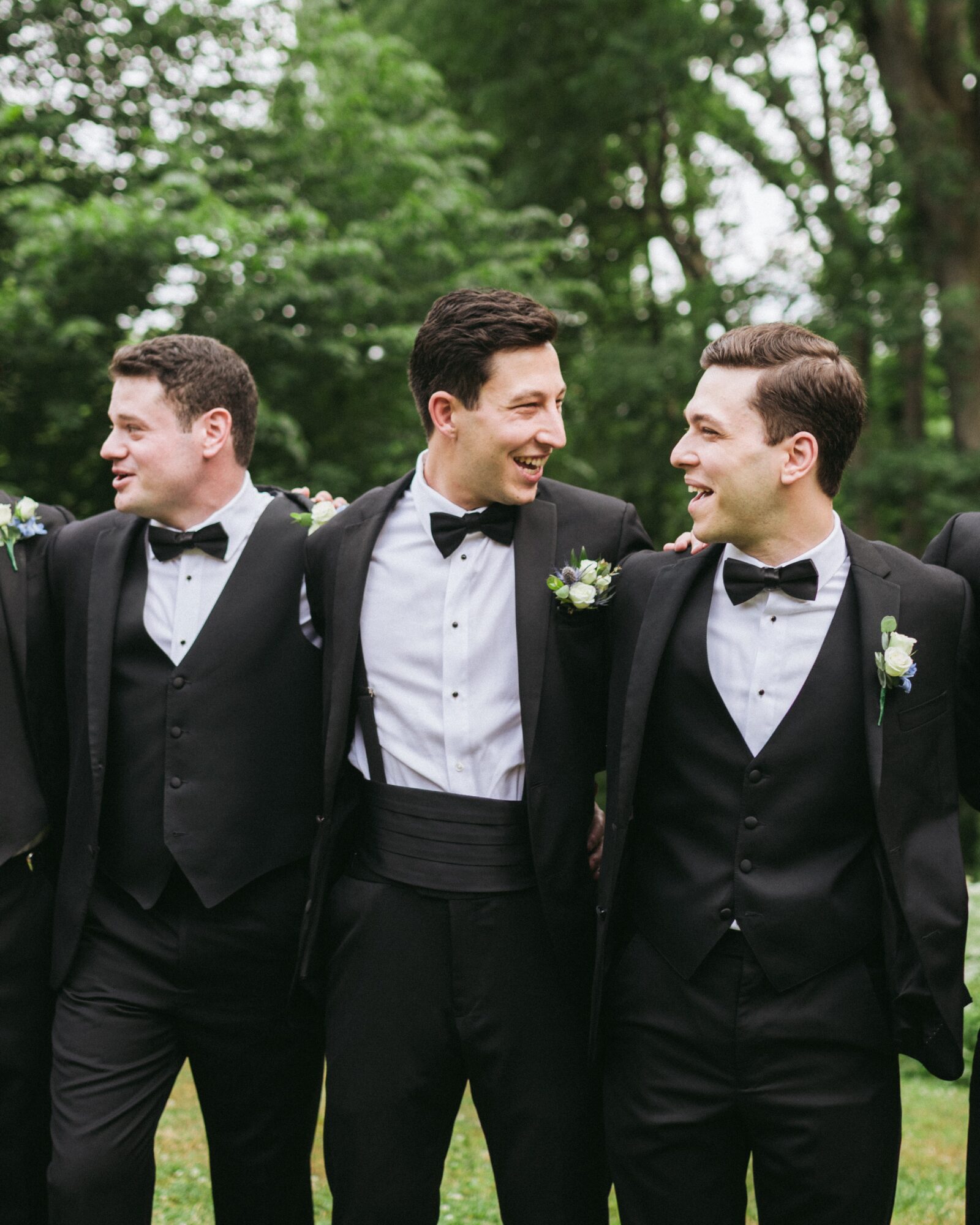 Three men in tuxedos smiling and talking outdoors with trees in the background.