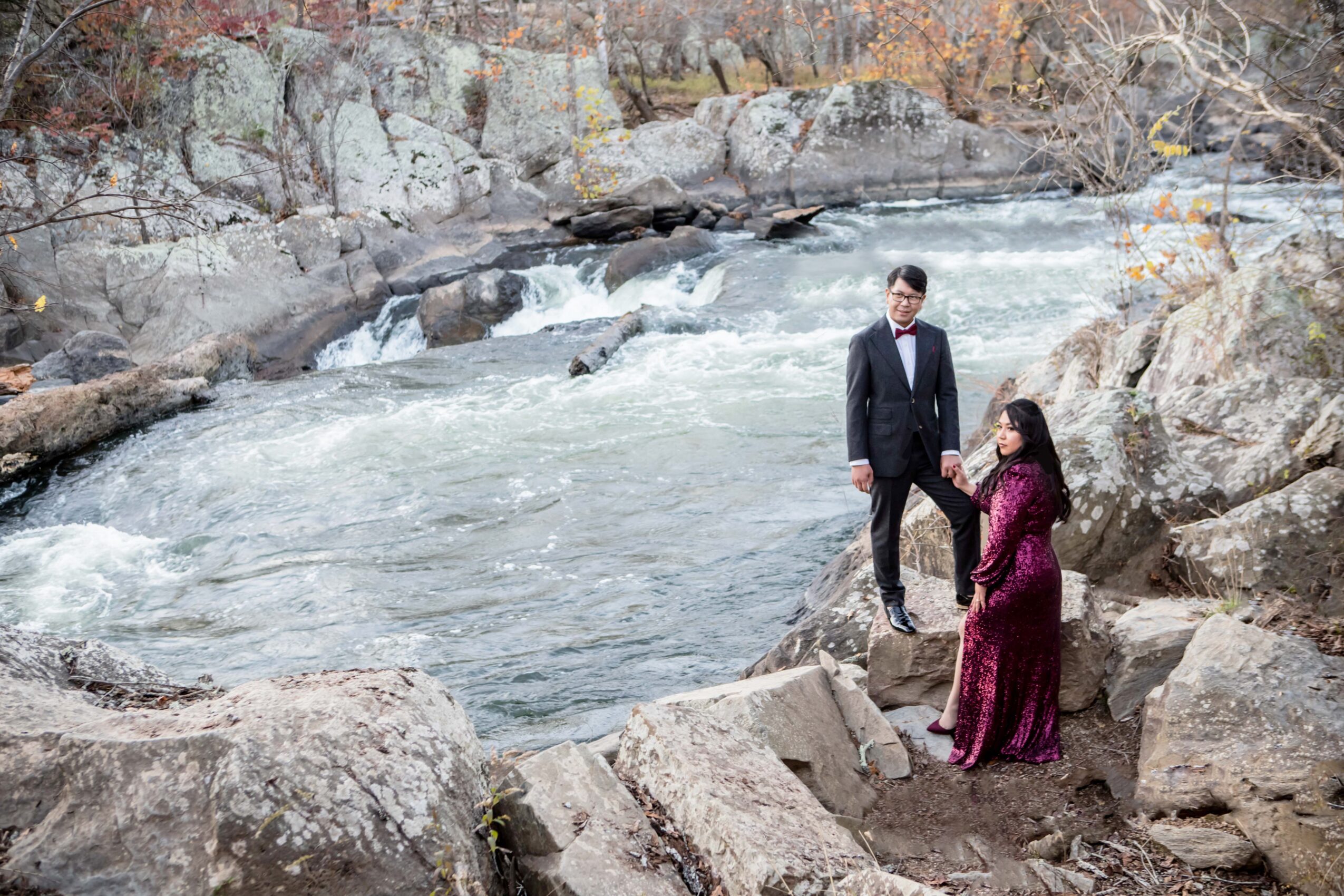 A man in a suit and a woman in a dress stand on rocks near a river with a small waterfall, surrounded by rocks and trees.
