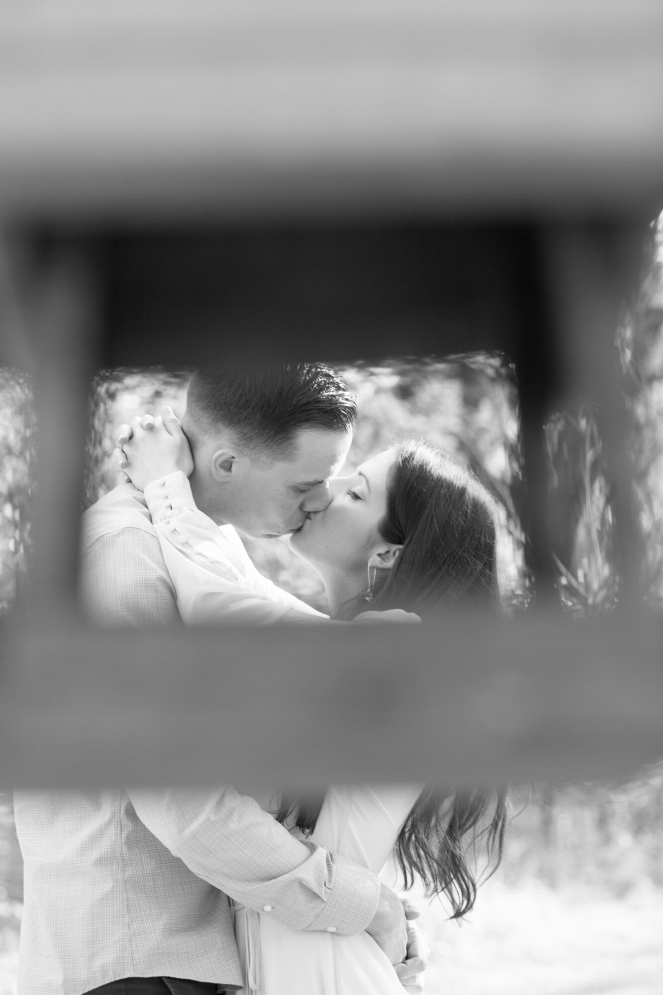 A couple kissing outdoors, seen through a wooden structure, in black and white.