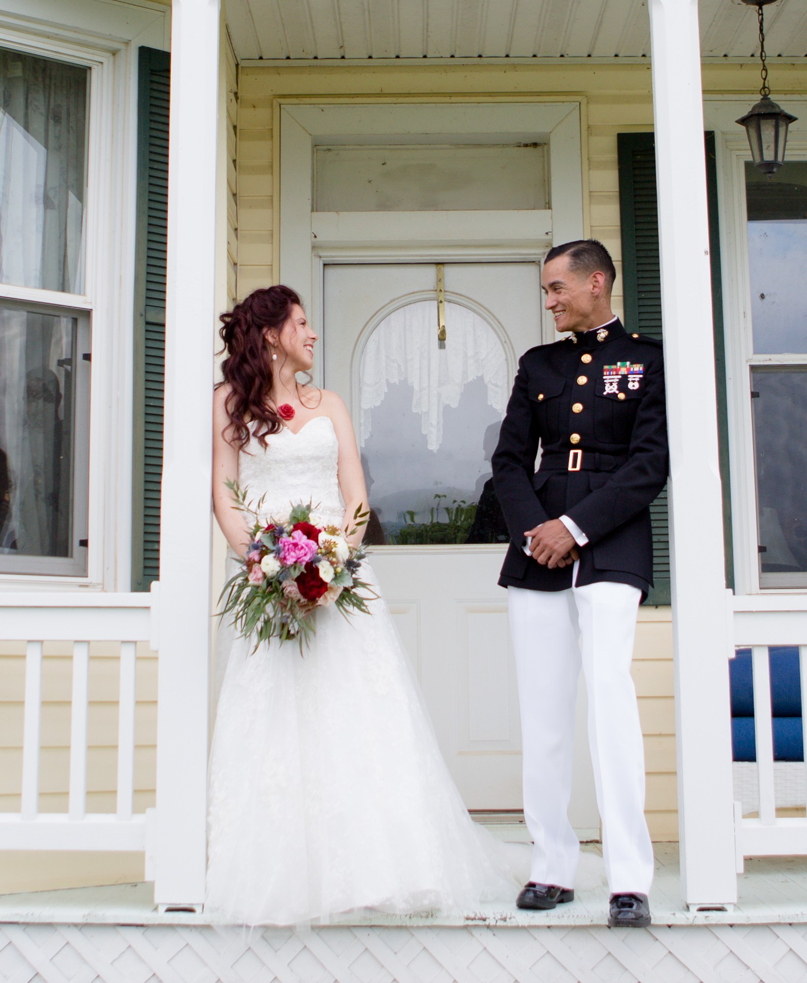 Bride and groom stand on porch, facing each other, smiling, with house door and windows in background.