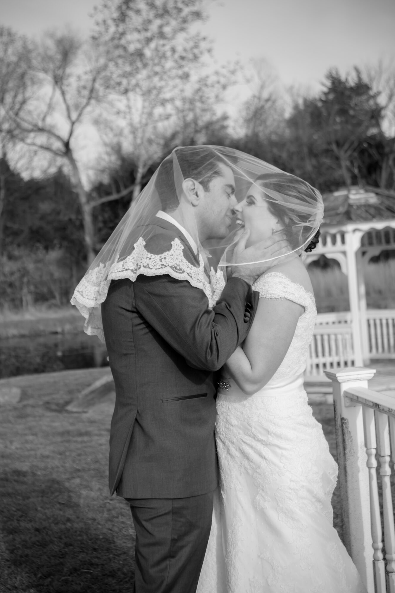 Couple in wedding attire under a veil, standing close outdoors with trees and a gazebo in background.