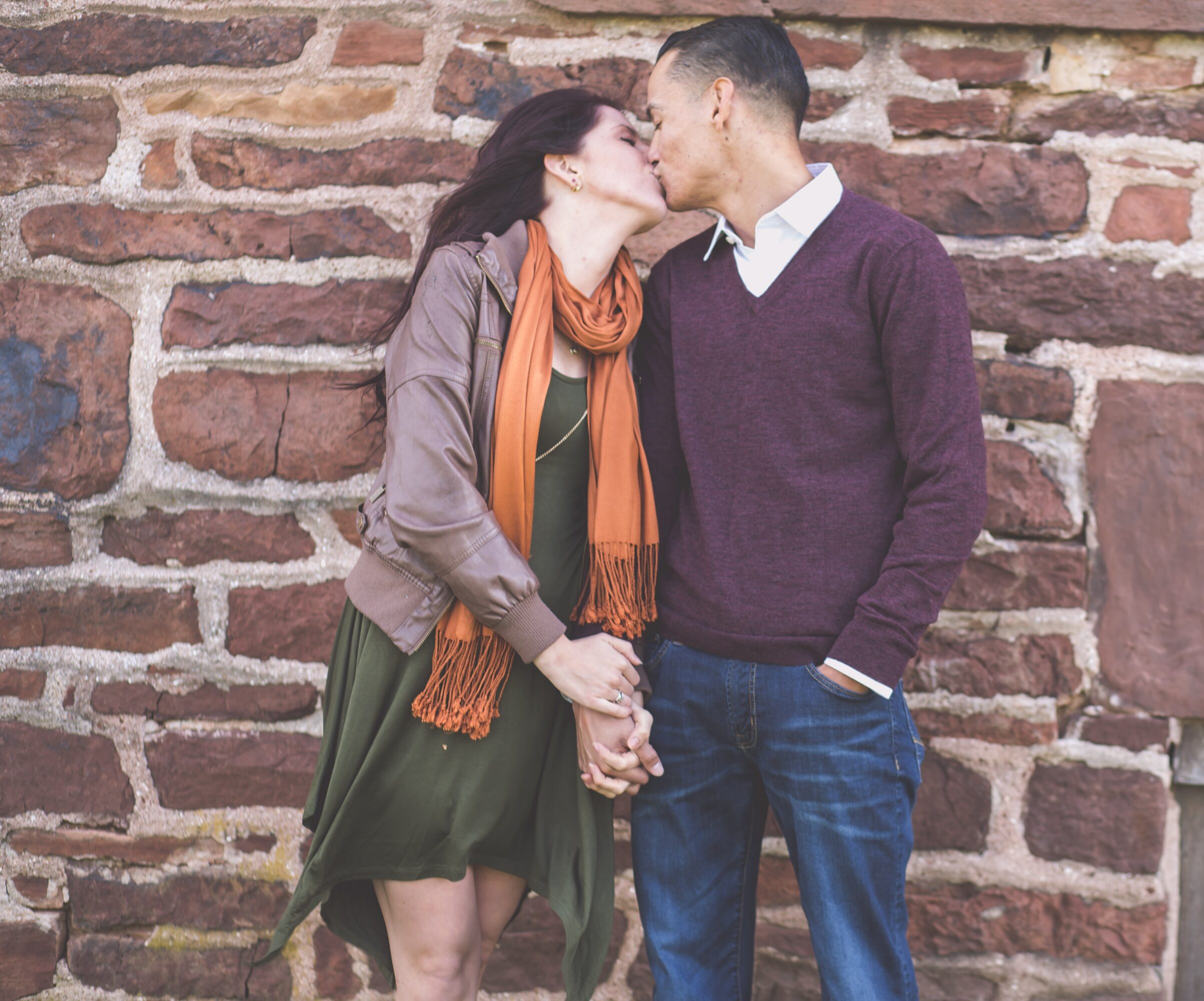 A couple stands close, holding hands, about to kiss against a brick wall background.