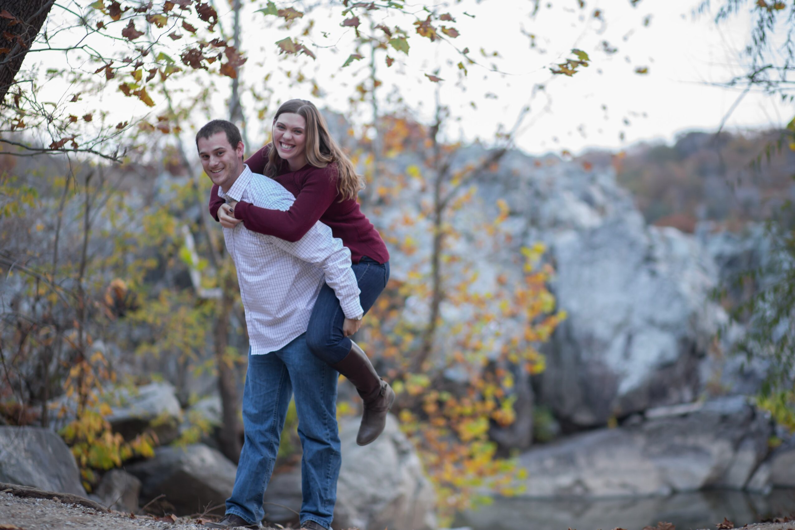 A couple outdoors, the woman on the man's back, surrounded by trees with autumn leaves and rocks.