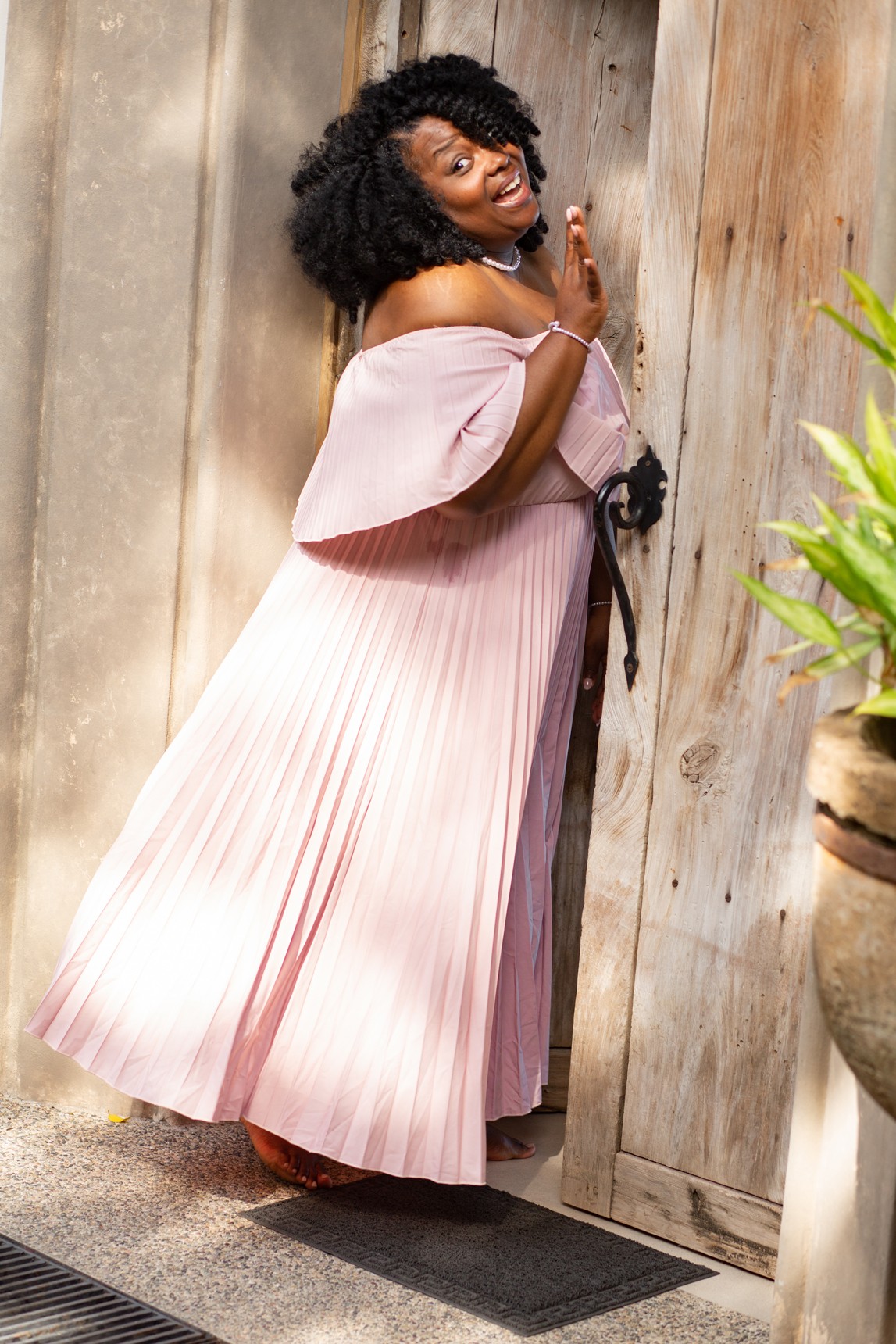 Woman with curly hair in a pink pleated dress standing by a wooden door, holding her finger to her lips.