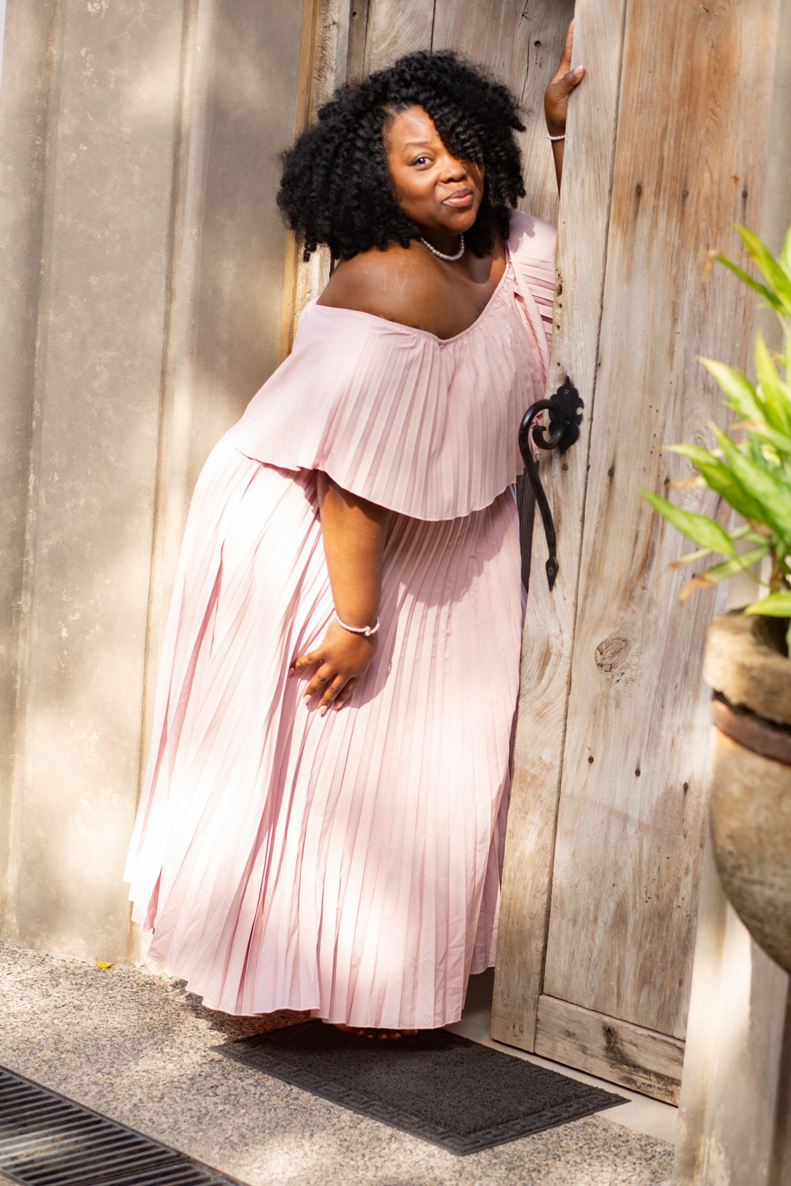 Woman with curly hair in a pink off-shoulder dress standing next to a wooden door, holding the door handle, outdoors.