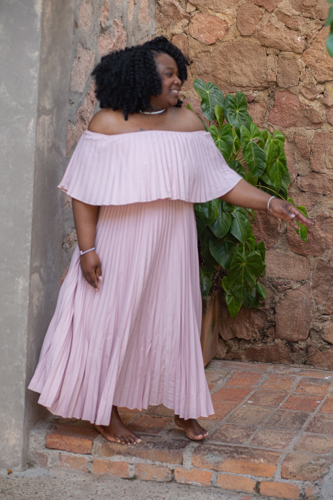 Woman in pink off-shoulder dress standing outdoors near a brick wall and green plant, smiling and pointing.