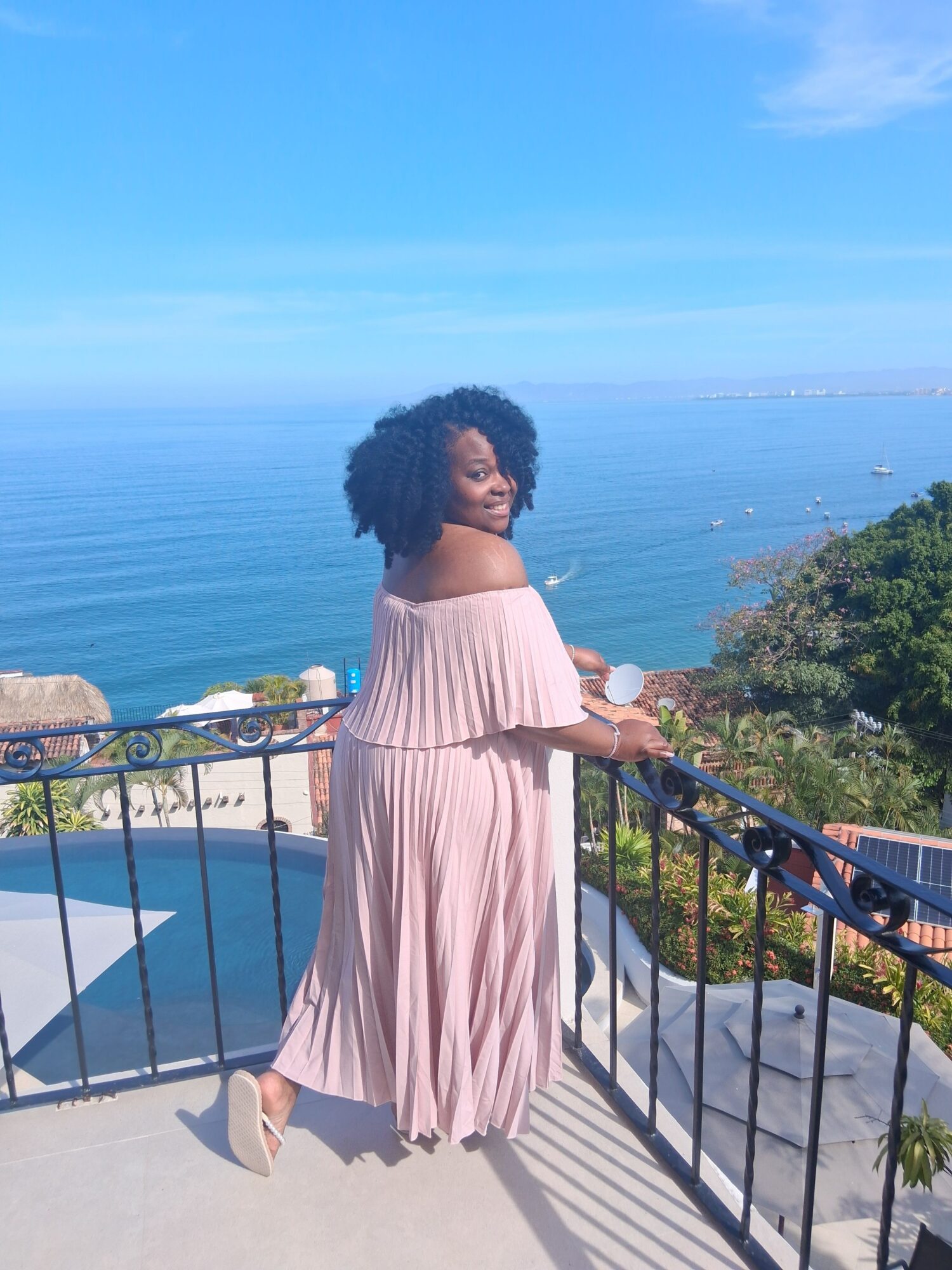 Woman in pink dress standing on a balcony overlooking water, trees, and boats, with a railing and clear sky.