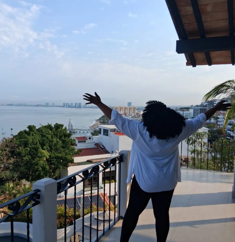 Person with arms outstretched on balcony overlooking city and water, with trees and buildings in background.