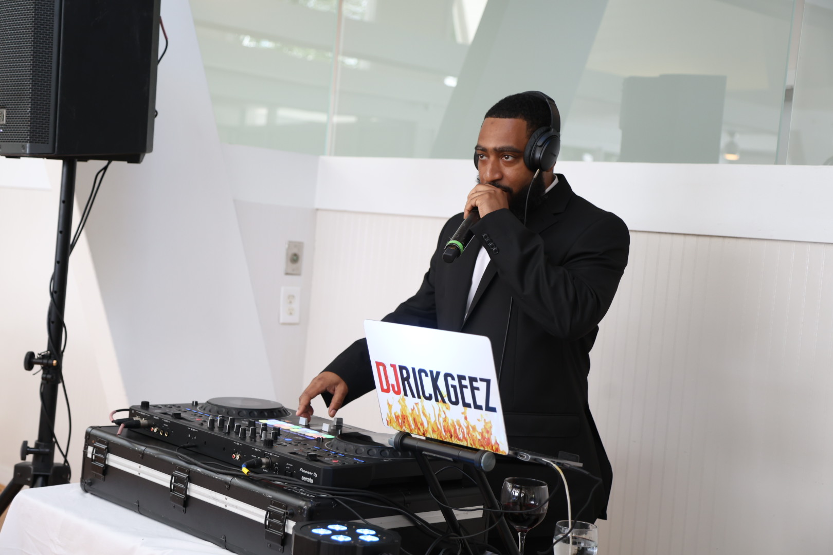 DJ wearing headphones and a suit, standing behind a DJ setup with a laptop, in a modern indoor space.