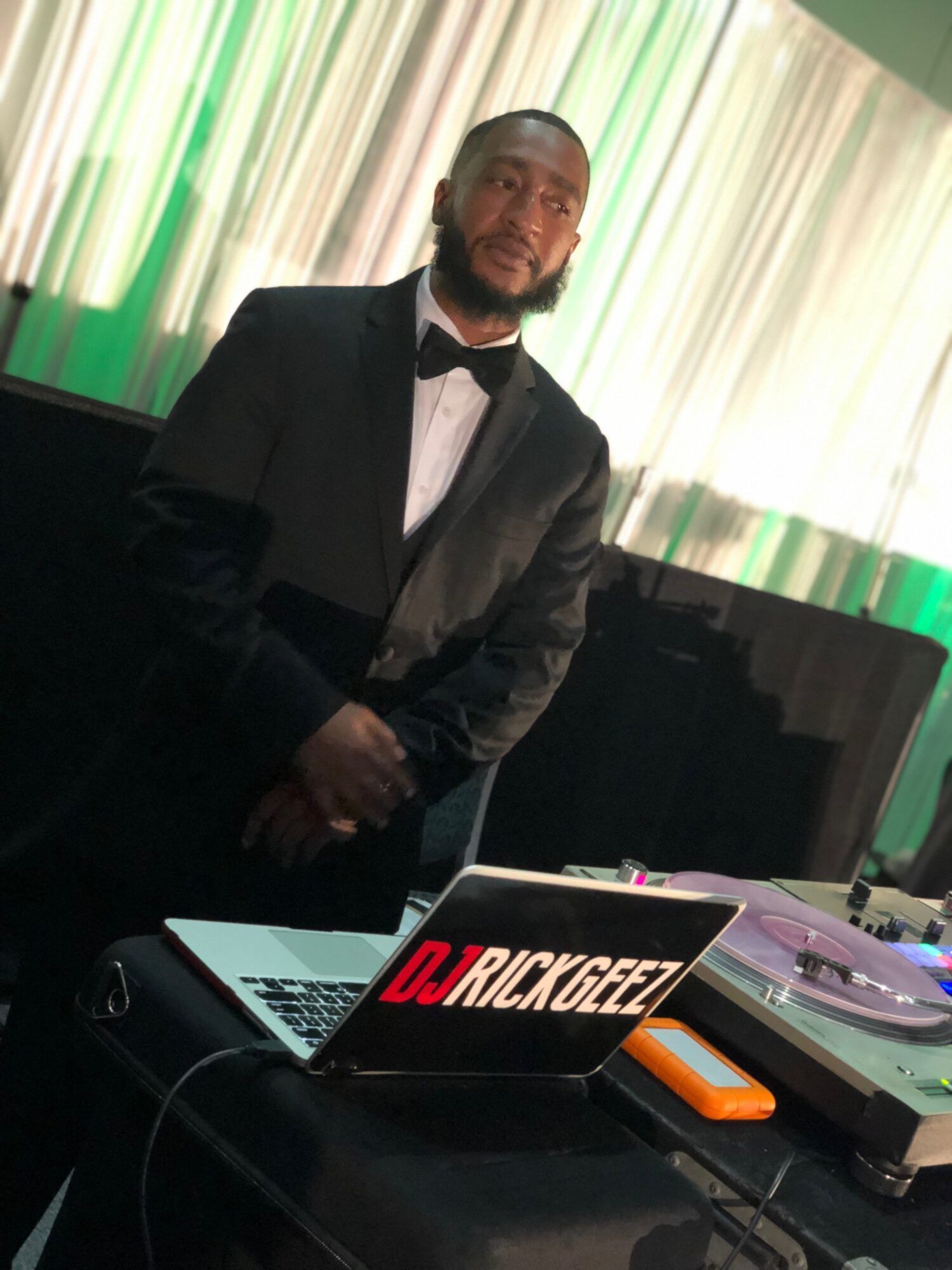 Man in tuxedo standing behind DJ equipment with a laptop, in front of a curtain with green and white lighting.