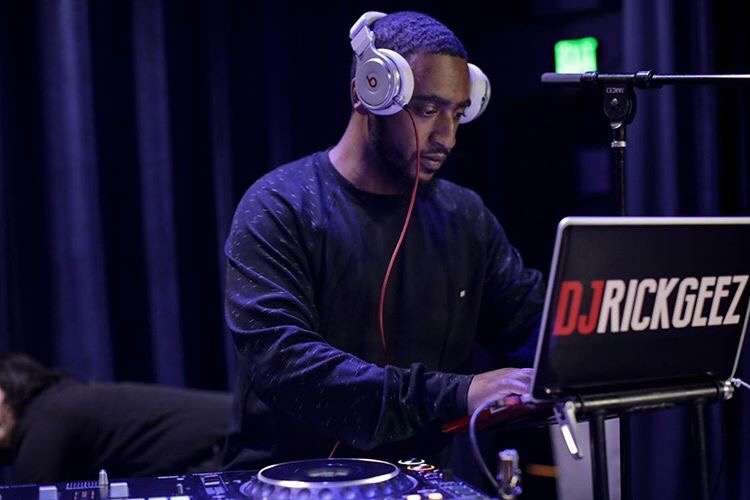 DJ wearing headphones working at a mixing console with a laptop displaying 'DJ RICKGEEZ' in a dark room