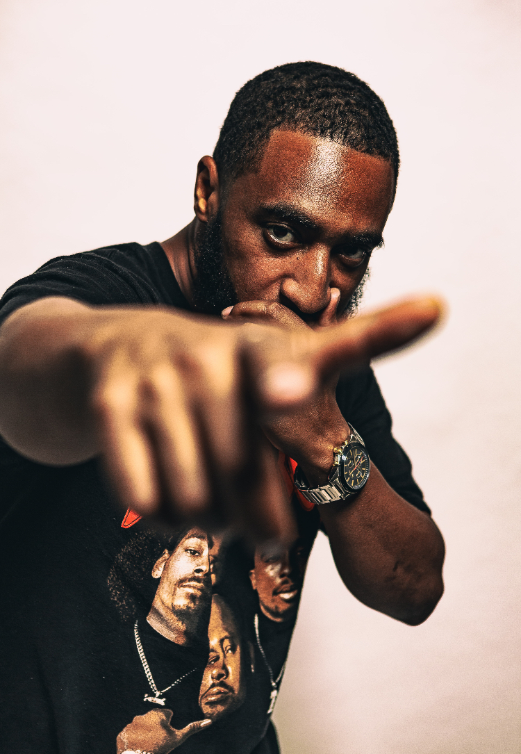 Man pointing directly at the camera, wearing a black T-shirt with images of people, against a plain background.