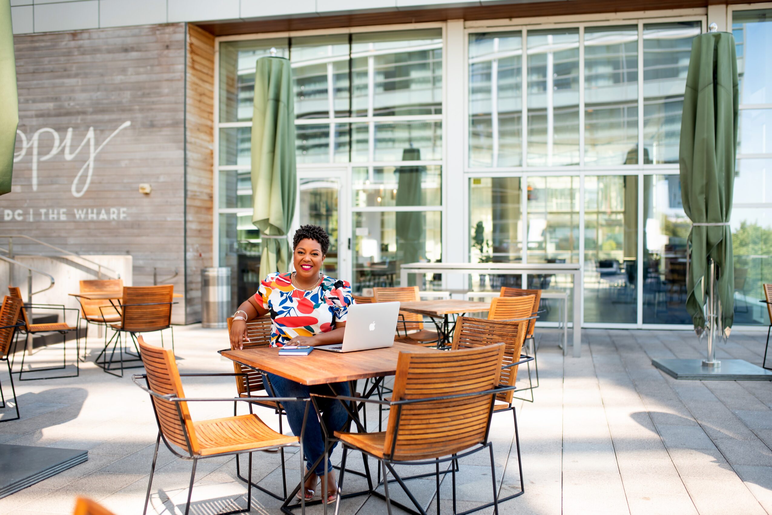 Woman sitting at outdoor table with laptop, smiling, in a modern patio area with large windows and green umbrellas.