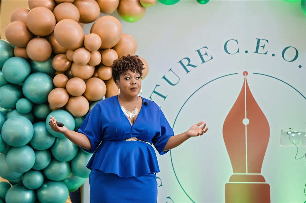 Woman in blue dress standing in front of a balloon backdrop and a logo with a pen nib symbol, gesturing with hands.