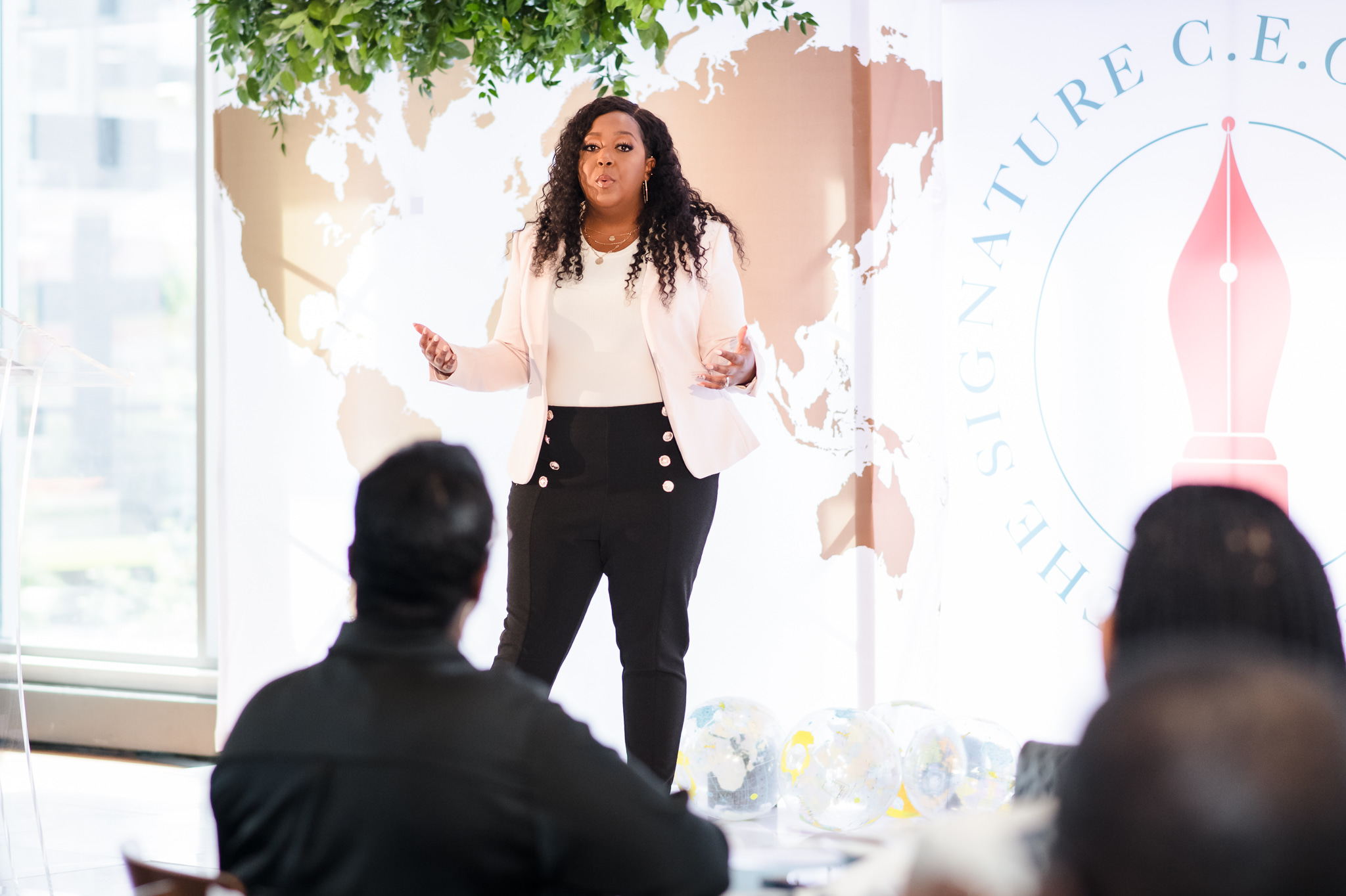 Woman speaking to an audience in a bright room with a world map and logo in the background.