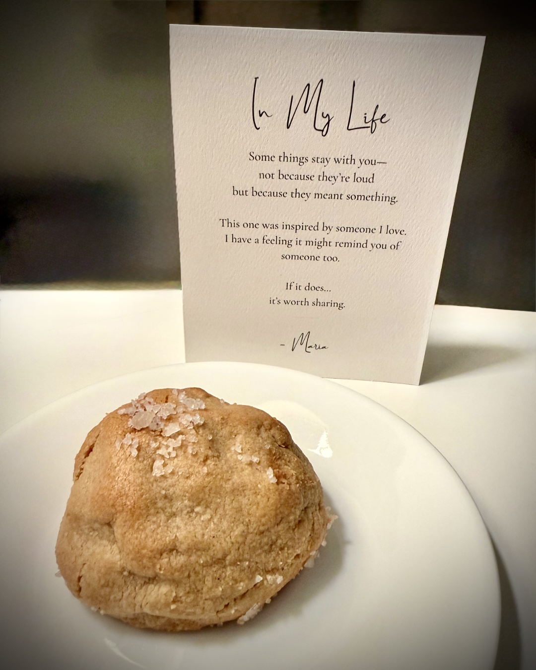 A cookie on a white plate with a note in the background, handwritten and decorated with small sugar crystals on the cookie.