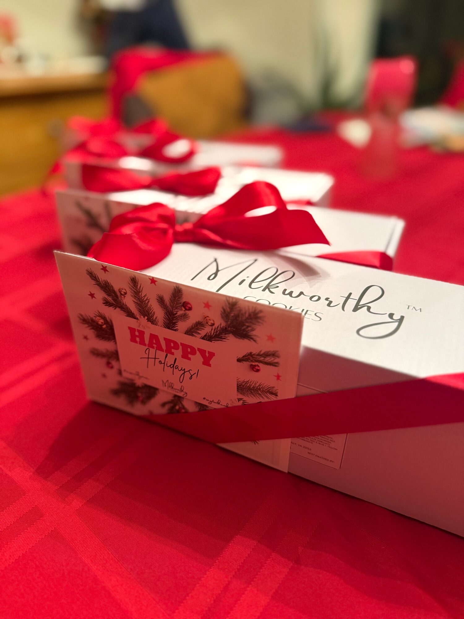 White gift boxes with red ribbons and a holiday card on a red tablecloth, background blurred.