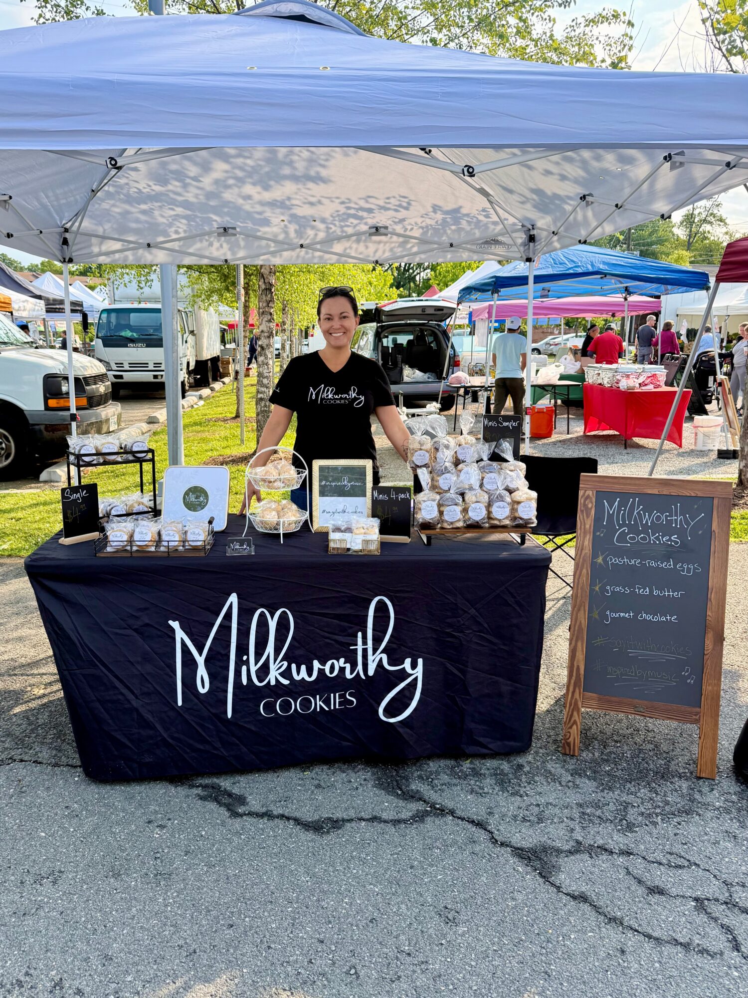 Vendor standing behind a table with cookies at an outdoor market, under a blue canopy, with other stalls in background.