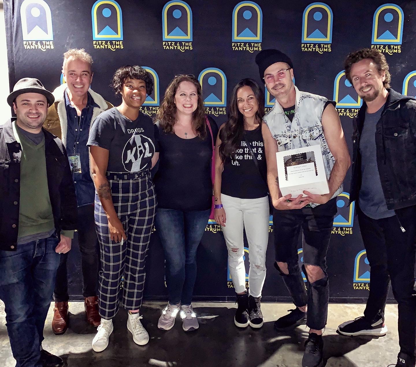 Group of seven people standing together in front of a backdrop with logos, smiling, some holding a box.