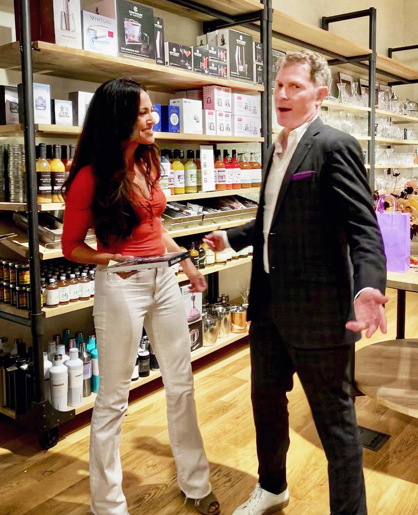 Two people talking in a store aisle with shelves of products behind them, one woman and one man, smiling and gesturing.