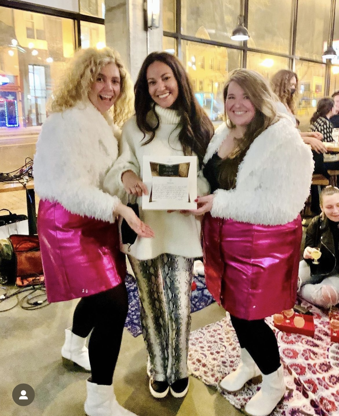 Three women standing indoors, smiling, holding a box, with a patterned rug and people in the background.