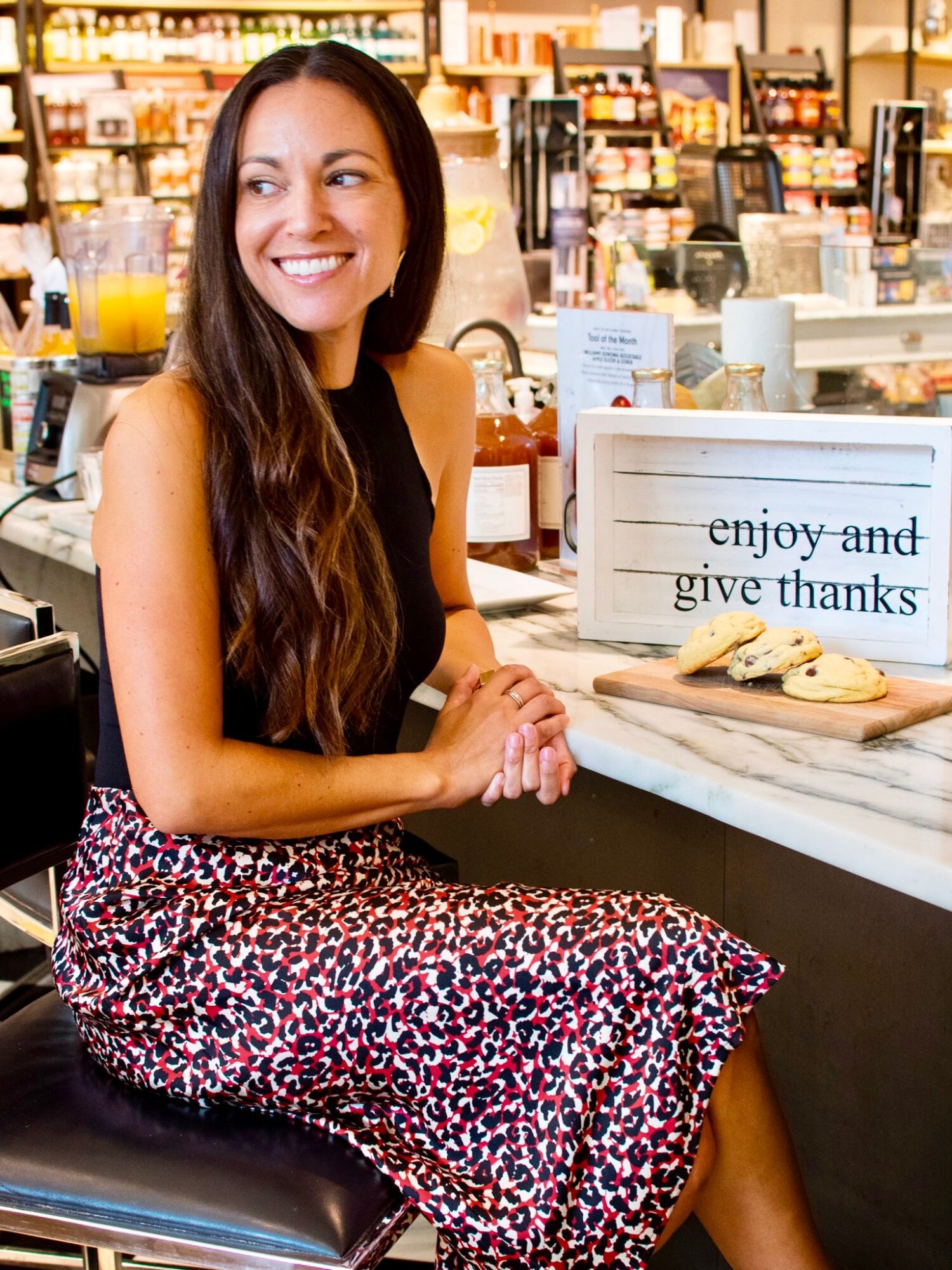 Woman with long hair smiling at a counter in a store, with a sign that says 'enjoy and give thanks', and cookies on the counter.