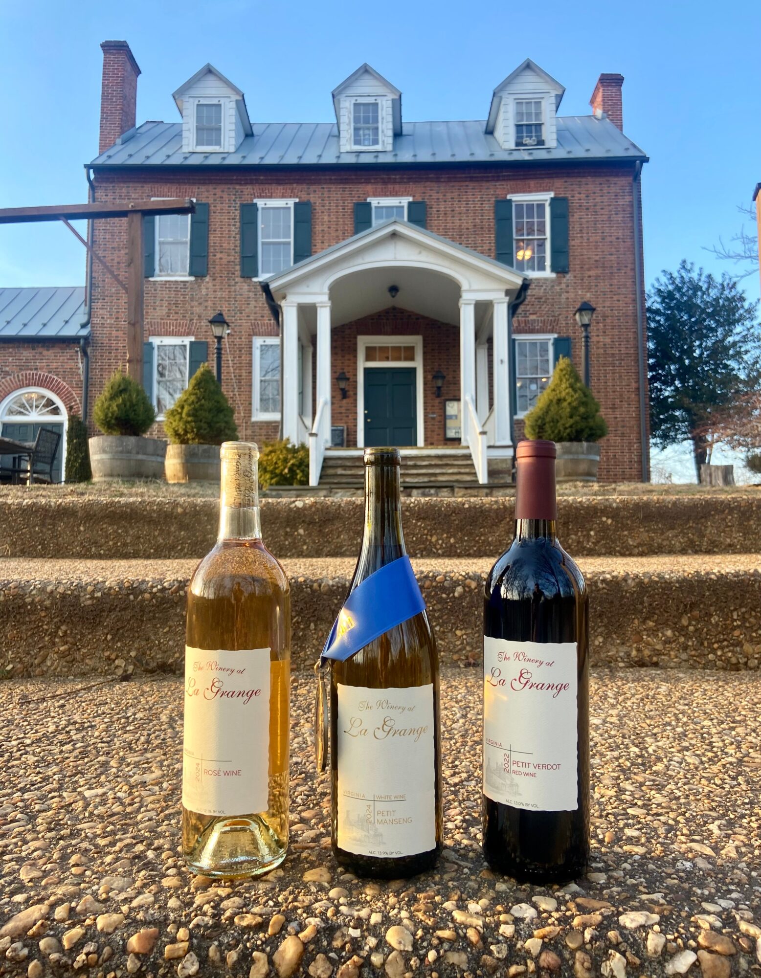 Three bottles of wine on steps in front of a large brick house with a porch and dormer windows.