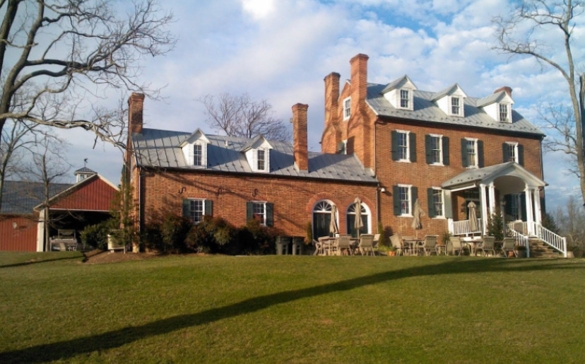 Large brick house with multiple chimneys, dormer windows, and a porch, surrounded by trees and a grassy lawn.