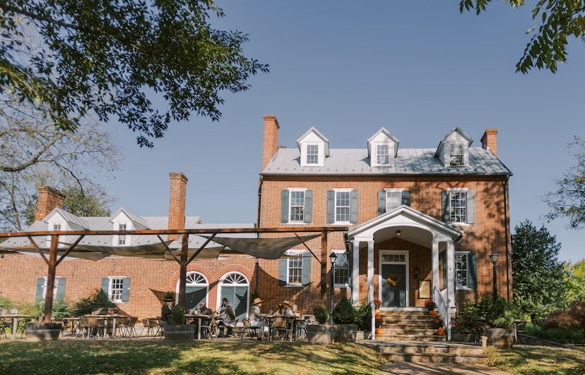 Large brick house with white porch, surrounded by trees and outdoor furniture, under a clear blue sky.