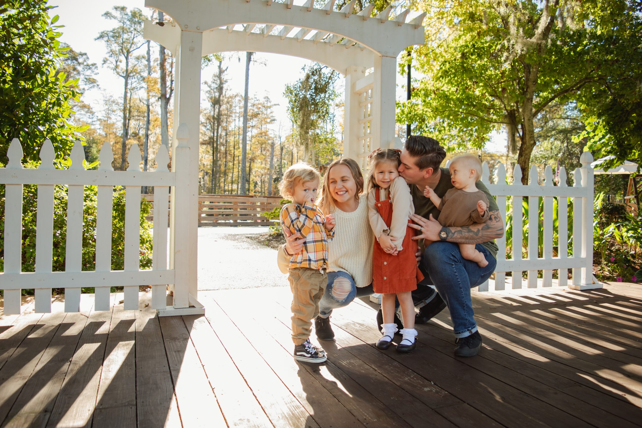 Group of children and a woman standing near a white garden gate outdoors, surrounded by trees and sunlight.