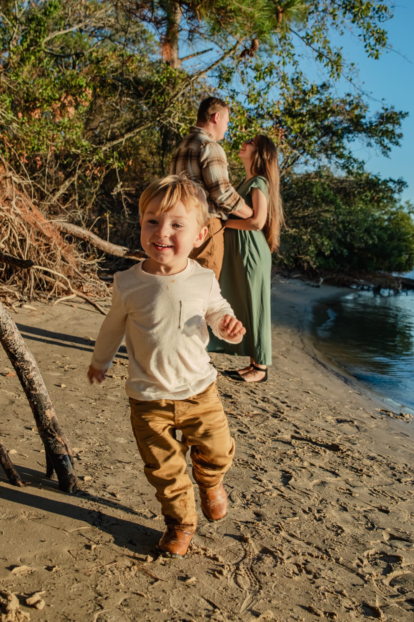 Young boy running on sandy beach near water with two women in the background, trees, and clear sky.