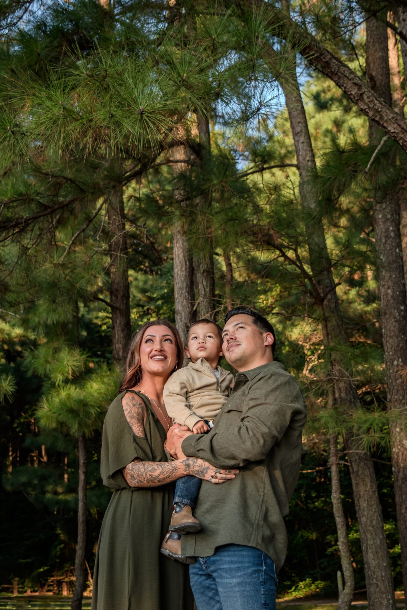 Family of three standing outdoors among tall trees, smiling and embracing each other, sunlight filtering through leaves.