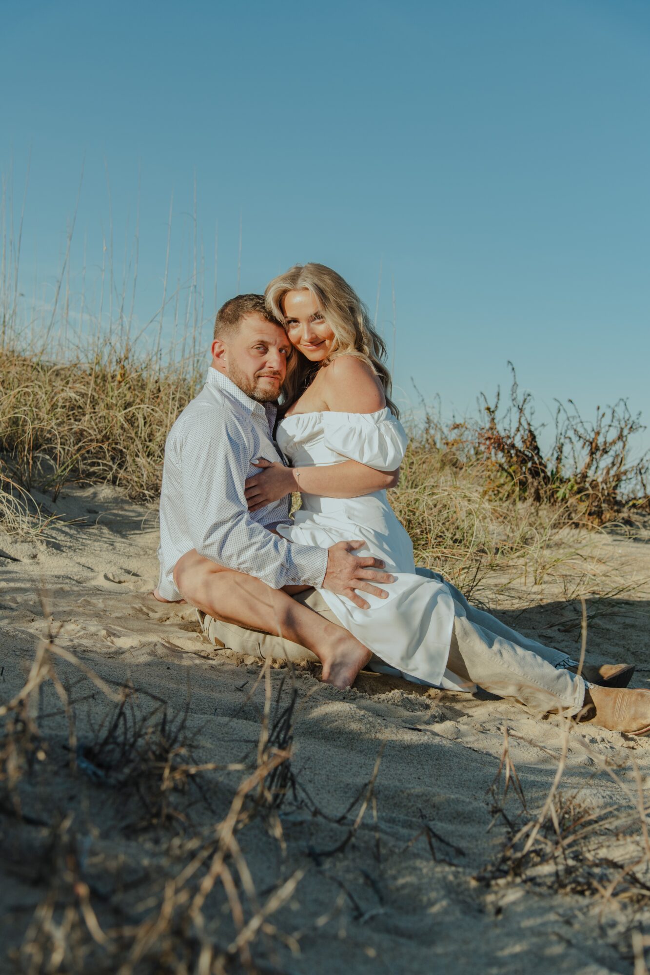 A couple sitting on a sandy beach, embracing each other, with clear blue sky and grassy dunes in the background.